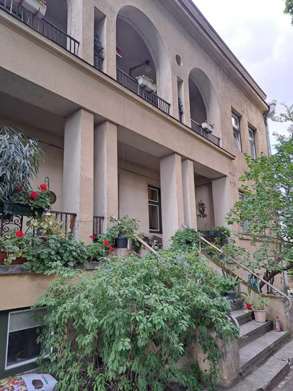 Didelis pastatas su arkinėmis nišomis, kolonomis ir daugybe gėlių vazonų prie įėjimo.