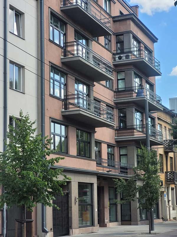 Rožinės spalvos daugiaaukštis pastatas su balkonais ir metaliniais turėklais.