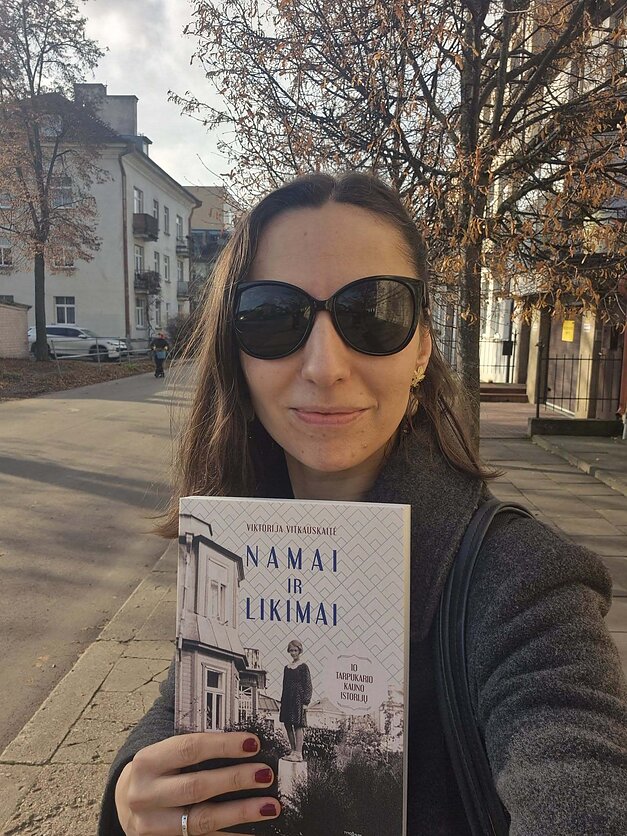 Viktorija stovi gatvėje su nuo saulės akiniais ir rankoje laiko savo knygą "Namai ir likimai"